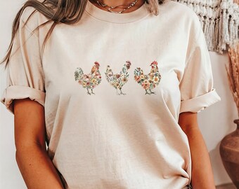 May include: A beige t-shirt with a floral design of three chickens. The chickens are facing forward and have a light brown, pink, and green floral pattern.