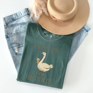 May include: A teal green t-shirt with a white goose holding a cup of coffee. The text "I run on honkin'" is printed on the shirt in brown.