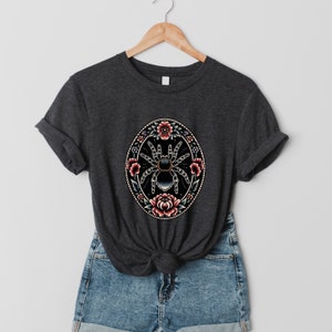 May include: A dark grey t-shirt with a black and blue spider surrounded by pink and red flowers in a circular design.