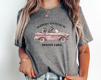 May include: A gray t-shirt with a graphic of two raccoons driving a pink car. The text "Support Your Local Street Cats" is printed on the shirt.