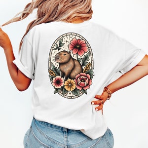 K&ouml;nnte beinhalten: Wei&szlig;es T-Shirt mit einer Vintage-Illustration eines Capybaras, umgeben von Blumen in einem runden Rahmen. Das Capybara ist braun und hat eine rosa Nase. Die Blumen sind rosa, gelb und rot.