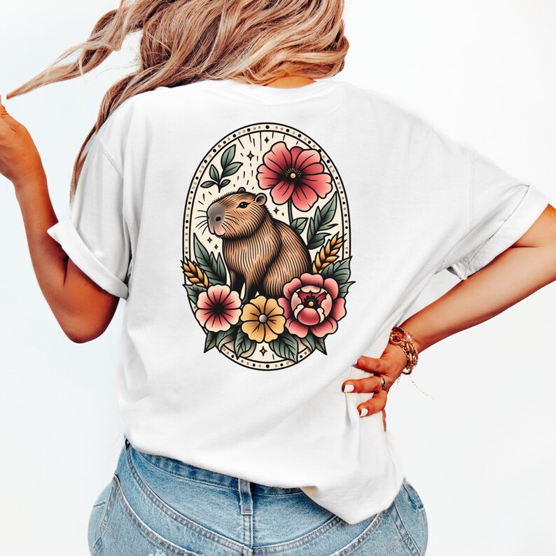 K&ouml;nnte beinhalten: Wei&szlig;es T-Shirt mit einer Vintage-Illustration eines Capybaras, umgeben von Blumen in einem runden Rahmen. Das Capybara ist braun und hat eine rosa Nase. Die Blumen sind rosa, gelb und rot.