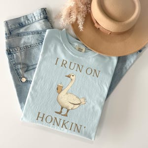 May include: A light blue t-shirt with the text "I run on honkin'" printed in brown. A cartoon goose holding a cup of coffee is printed on the shirt.