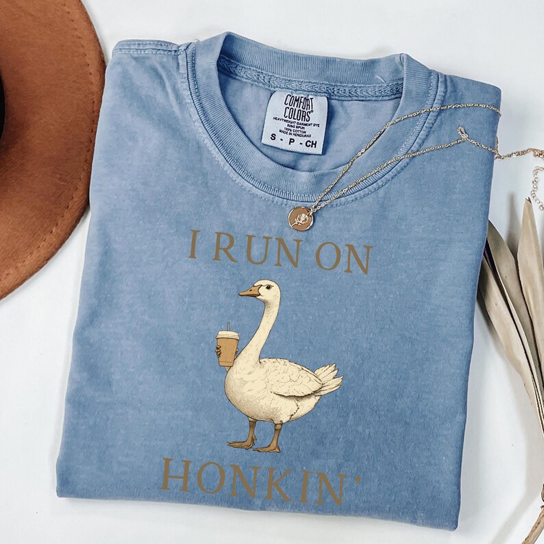 May include: A light blue t-shirt with a distressed look features a cartoon goose holding a cup of coffee. The text "I run on honkin'" is printed in brown on the shirt.