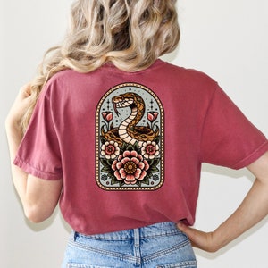 May include: A maroon t-shirt with a vintage-style illustration of a snake coiled around flowers. The illustration is framed in a light blue arch with a white background.