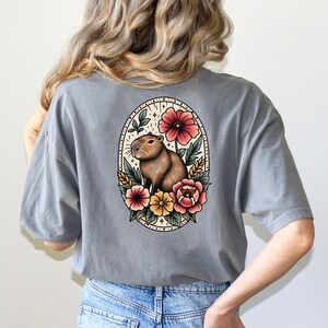 K&ouml;nnte beinhalten: Ein graues T-Shirt mit einer Vintage-Illustration eines Capybaras, umgeben von Blumen. Das Capybara ist braun und wei&szlig;, und die Blumen sind rot, rosa, gelb und wei&szlig;.