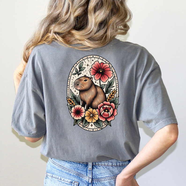 K&ouml;nnte beinhalten: Ein graues T-Shirt mit einer Vintage-Illustration eines Capybaras, umgeben von Blumen. Das Capybara ist braun und wei&szlig;, und die Blumen sind rot, rosa, gelb und wei&szlig;.