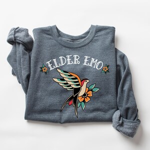 May include: A dark gray sweatshirt with the text "ELDER EMO" in white lettering. A stylized tattoo-style bird with colorful wings and a flower is centered on the shirt.