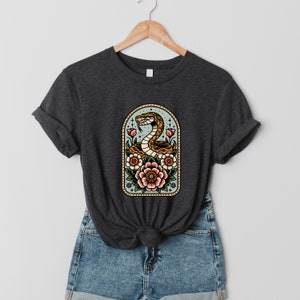 Könnte beinhalten: Ein dunkelgraues T-Shirt mit einer Vintage-Illustration einer Schlange, die sich um Blumen in einem goldenen Rahmen windet. Die Blumen sind rosa, rot und weiß.