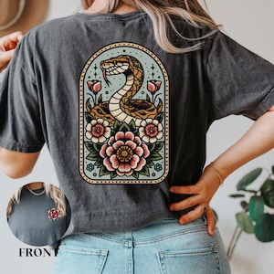 May include: A dark grey t-shirt featuring a vintage-style illustration of a snake entwined with flowers. The snake is yellow and brown, and the flowers are pink, red, and blue. The illustration is enclosed within a light blue oval with a gold border.