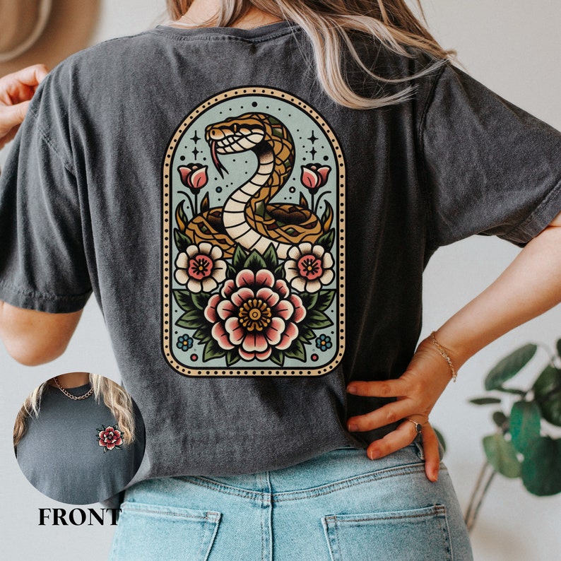 May include: A dark gray t-shirt with a vintage-style illustration of a snake wrapped around flowers. The snake is yellow and brown, and the flowers are pink, red, and blue. The illustration is surrounded by a light blue oval with a gold border.