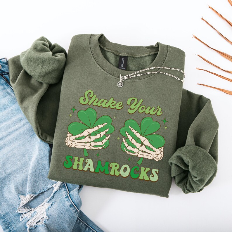 Puede incluir: Sudadera verde con una mano de esqueleto sosteniendo un tr&eacute;bol y el texto "Shake Your Shamrocks".