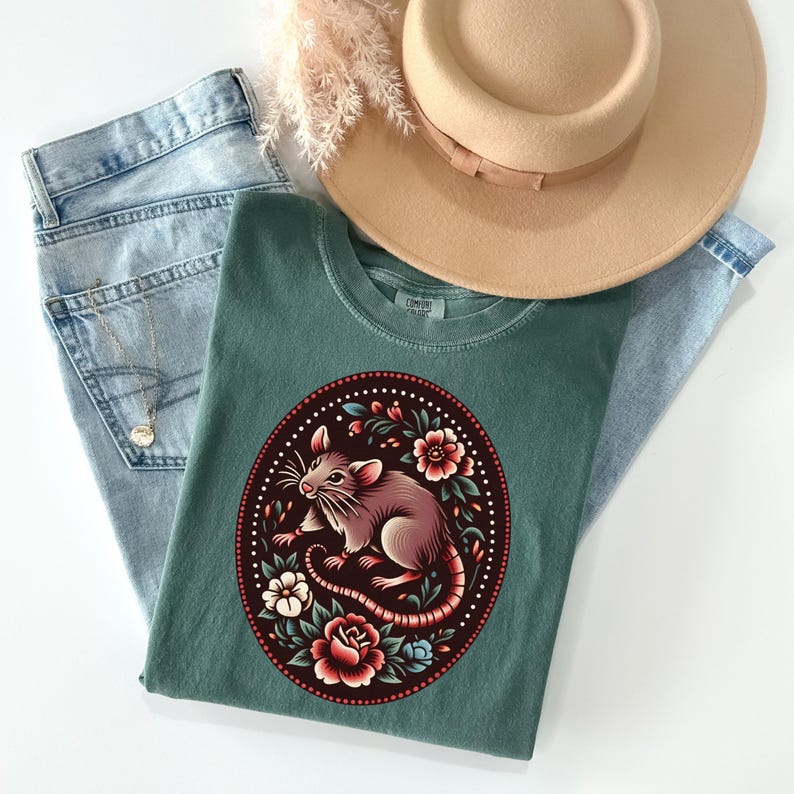 K&ouml;nnte beinhalten: Ein tealgr&uuml;nes T-Shirt mit einem floralen Design, in dessen Mitte sich eine Ratte befindet. Die Ratte ist braun und wei&szlig; mit einem langen Schwanz. Die Blumen sind rot, rosa und wei&szlig;.