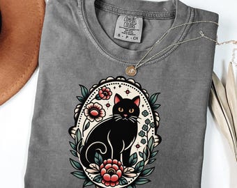 May include: A charcoal gray t-shirt featuring a graphic of a black cat centered within an oval frame decorated with red flowers and green leaves. The inner neck label displays the text Comfort Colors, 100% cotton, and size S P CH.