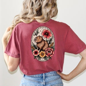 K&ouml;nnte beinhalten: Ein maroonfarbenes T-Shirt mit einer Vintage-Illustration eines Capybaras, umgeben von Blumen. Die Illustration befindet sich in einem ovalen Rahmen mit einem floralen Rand.