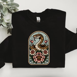 Peut inclure: Sweat-shirt noir avec une illustration de style vintage d'un serpent enroulé autour de fleurs dans un cadre bleu et vert. Les fleurs sont roses, rouges et jaunes.