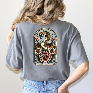 May include: A gray t-shirt with a vintage-style illustration of a snake wrapped around flowers. The snake is brown and yellow, and the flowers are pink, red, and yellow. The illustration is surrounded by a light blue background.