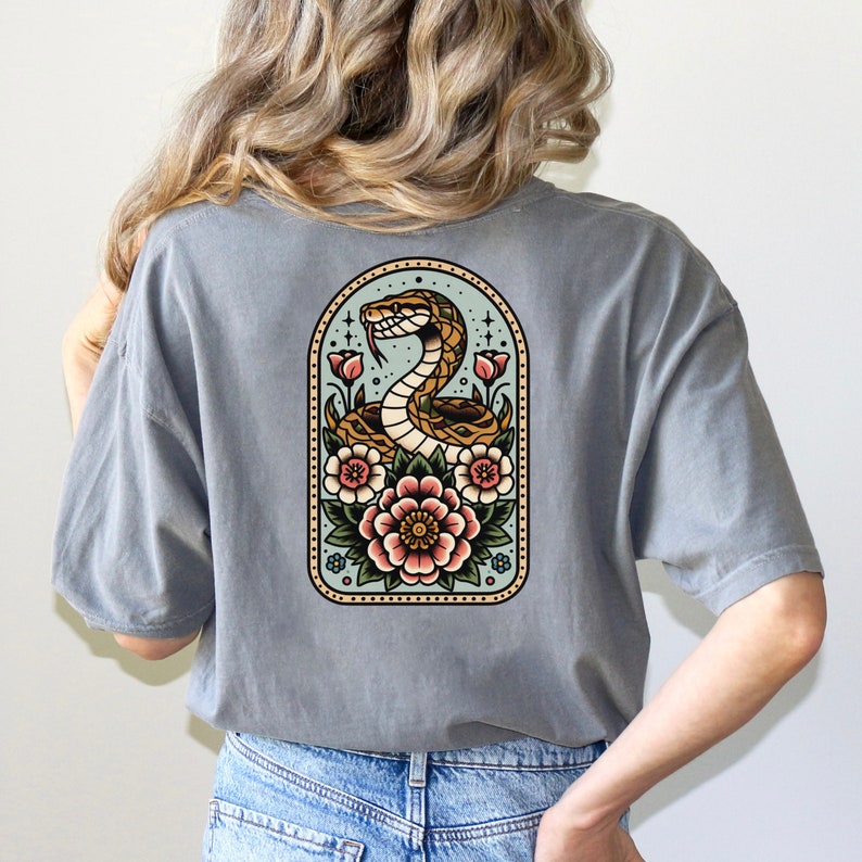 May include: A gray t-shirt with a vintage-style illustration of a snake wrapped around flowers. The snake is brown and yellow, and the flowers are pink, red, and yellow. The illustration is surrounded by a light blue background.