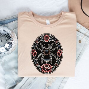 May include: A beige t-shirt with a black and gray tarantula spider surrounded by colorful flowers in a circular design.