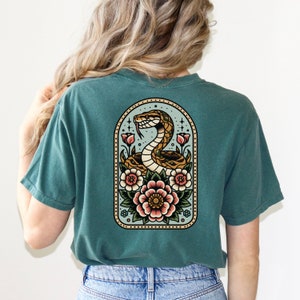 May include: A teal blue t-shirt with a vintage-style illustration of a snake coiled around flowers in a decorative frame. The snake is brown and yellow, and the flowers are pink, red, and yellow.