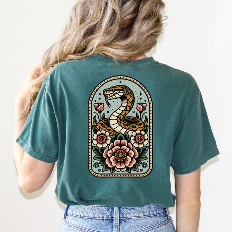 May include: A teal blue t-shirt with a vintage-style illustration of a snake coiled around flowers in a decorative frame. The snake is brown and yellow, and the flowers are pink, red, and yellow.