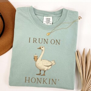 May include: A light blue t-shirt with a cartoon goose holding a coffee cup and the text "I run on honkin'" printed on it.