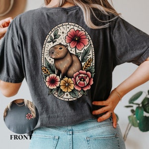 K&ouml;nnte beinhalten: Ein dunkelgraues T-Shirt mit einer Vintage-Illustration eines Capybaras, umgeben von Blumen. Die Illustration befindet sich in einem ovalen Rahmen mit einem floralen Rand. Das Capybara ist braun und hat eine wei&szlig;e Nase. Die Blumen sind rot, rosa, gelb und wei&szlig;.