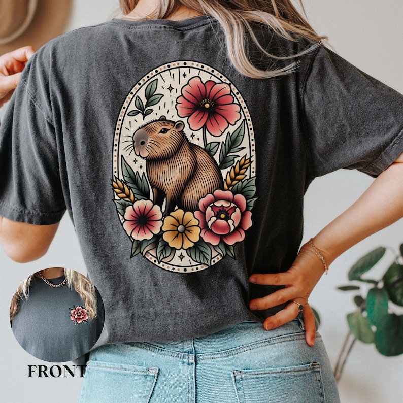 K&ouml;nnte beinhalten: Ein dunkelgraues T-Shirt mit einer Vintage-Illustration eines Capybaras, umgeben von Blumen. Die Illustration befindet sich in einem ovalen Rahmen mit einem floralen Rand. Das Capybara ist braun und hat eine wei&szlig;e Nase. Die Blumen sind rot, rosa, gelb und wei&szlig;.