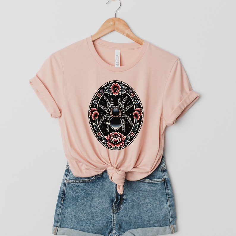 May include: A pink t-shirt with a black and white graphic of a spider surrounded by red and pink flowers. The graphic is in a circle with a black outline.
