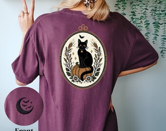 Könnte beinhalten: Ein maroonfarbenes T-Shirt mit einer schwarzen Katze, die auf einem Kürbis in einem goldenen Rahmen mit floralen Akzenten sitzt. Die Katze trägt ein goldenes Halsband und blickt nach links. Das T-Shirt hat auf der Vorderseite einen Halbmond und Fledermäuse.