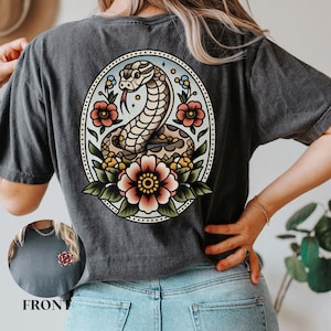 Puede incluir: Una camiseta gris oscuro con una ilustración de estilo vintage de una serpiente enroscada alrededor de flores. La serpiente es marrón y amarilla, y las flores son rojas, rosas y verdes. La ilustración está rodeada por un círculo con un borde punteado.