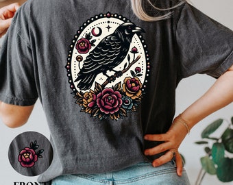 Puede incluir: Una camiseta gris oscuro con un cuervo negro posado en una rama con flores. El cuervo está rodeado de un círculo de flores en tonos rojos, amarillos y naranjas. El diseño es de estilo vintage.