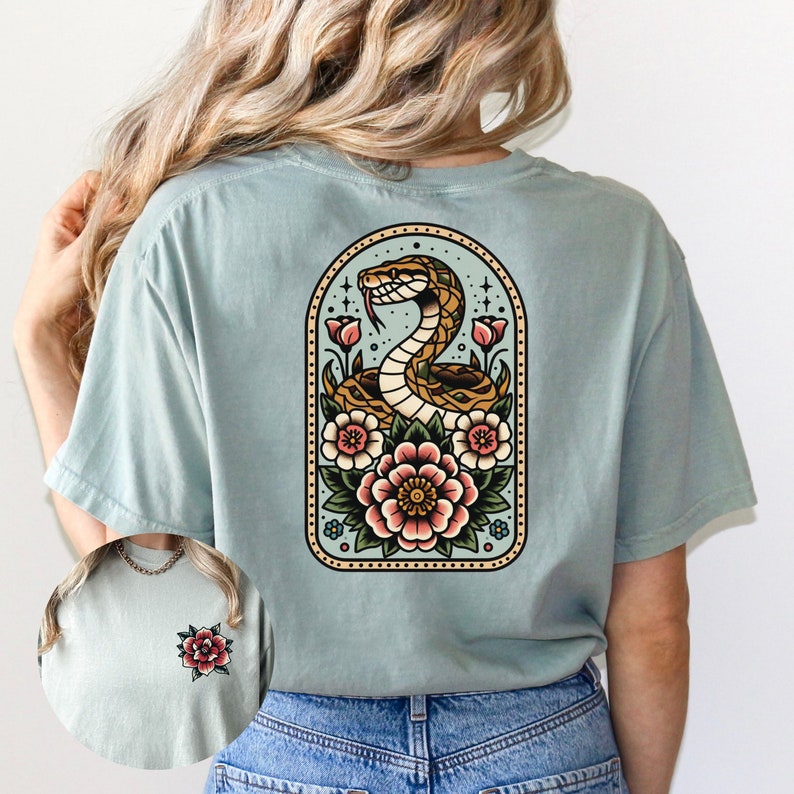 May include: A light blue t-shirt with a vintage-style illustration of a snake wrapped around flowers in a circular frame. The snake is brown and yellow, and the flowers are pink, red, and yellow.