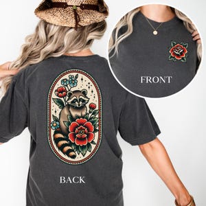 Könnte beinhalten: Ein anthrazitfarbenes T-Shirt mit einer Vintage-Illustration eines Waschbären auf der Rückseite. Der Waschbär ist von Blumen und Blättern in einem runden Rahmen umgeben. Die Vorderseite des Shirts hat eine einzelne rote Rose mit grünen Blättern.