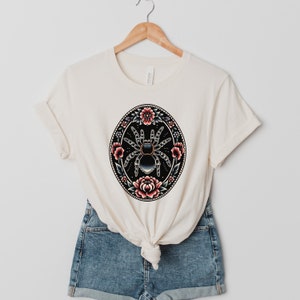 May include: A white t-shirt with a black and blue spider surrounded by pink and red flowers in a circular design.