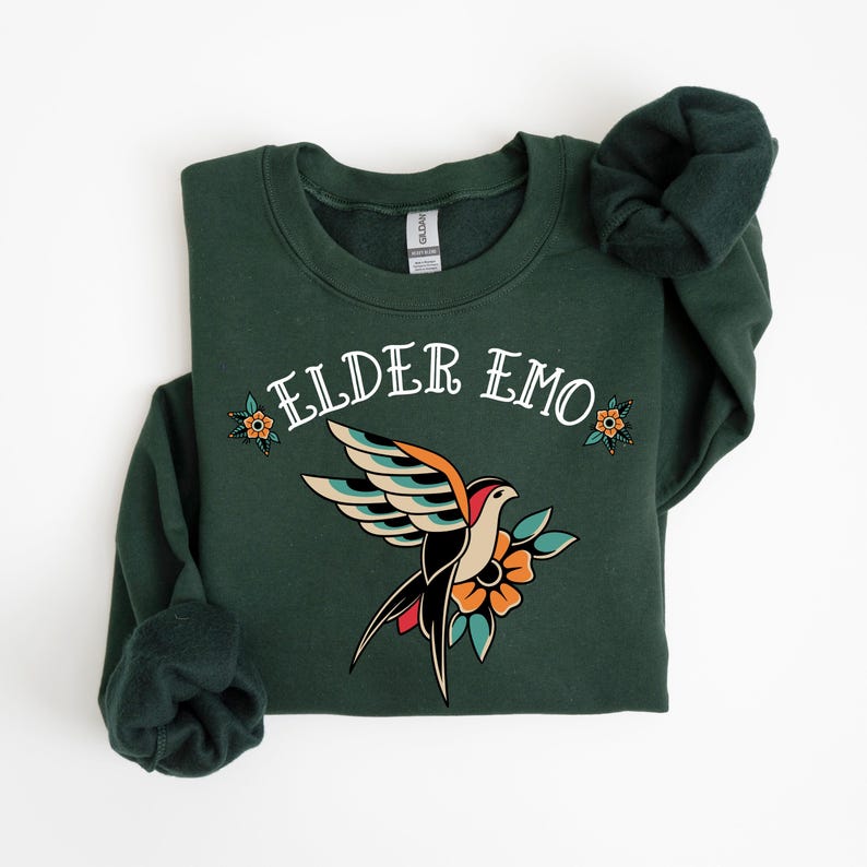 May include: A dark green sweatshirt with the text "ELDER EMO" in white lettering. A stylized tattoo-style bird with red, yellow, blue, and black accents is centered on the sweatshirt.