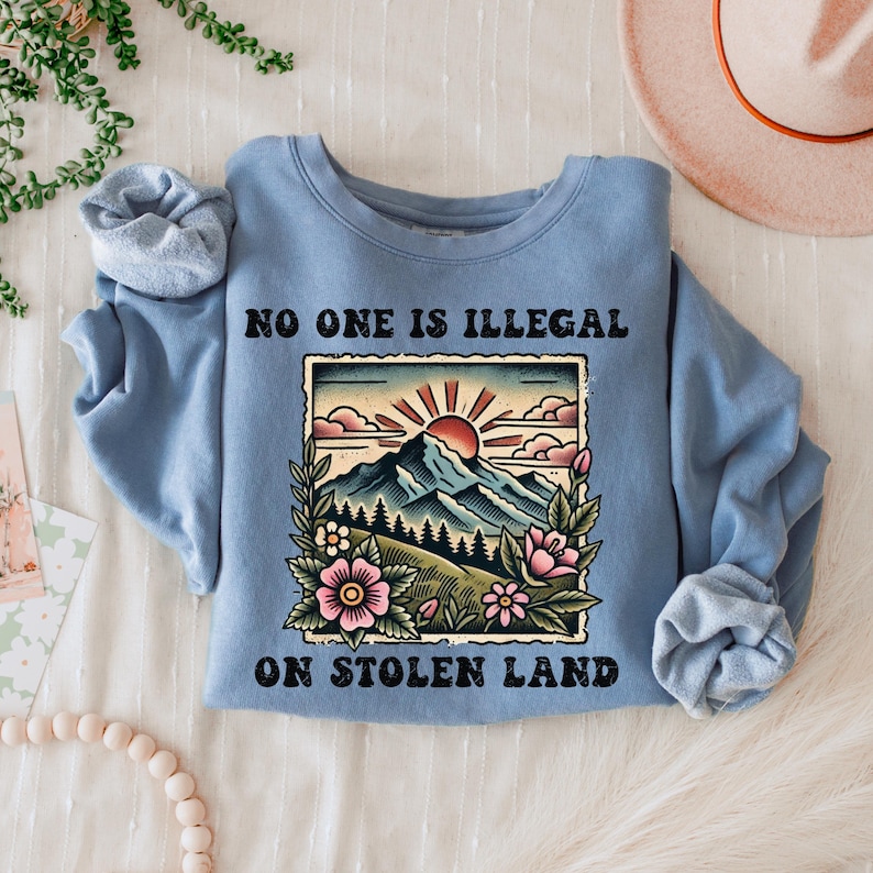May include: A light blue sweatshirt with the text "No one is illegal on stolen land" printed on the front. The text is in black and is surrounded by a vintage-style illustration of a mountain range with a sunset and flowers.