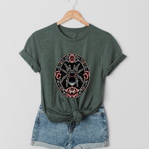 May include: A dark green t-shirt with a black and white spider surrounded by pink and red flowers in a circle.