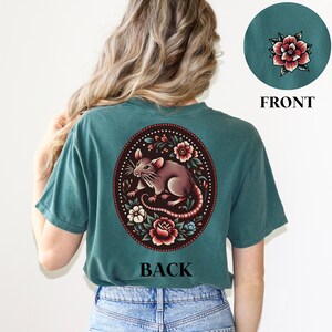 May include: Teal blue t-shirt with a floral design featuring a rat on the back. The design is in a circle with a white outline. The text "BACK" is printed below the design.