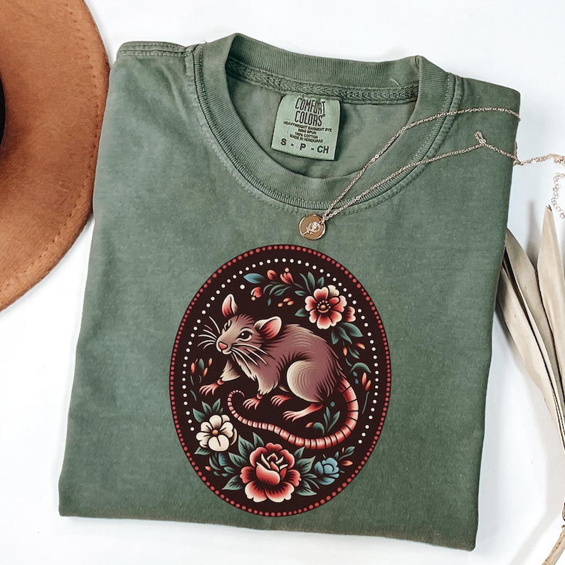 K&ouml;nnte beinhalten: Ein gr&uuml;nes T-Shirt mit einem floralen Design, das einen Ratten im Zentrum zeigt. Das Design ist in Rot-, Rosa- und Blaut&ouml;nen gehalten. Das Shirt hat ein Etikett mit der Aufschrift "Comfort Colors" und "S - P - CH".