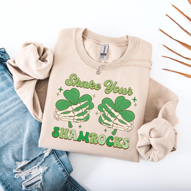 Puede incluir: Una sudadera de color beige con el texto "Shake Your Shamrocks" en letras verdes y blancas. Dos manos esquel&eacute;ticas sostienen tr&eacute;boles verdes.