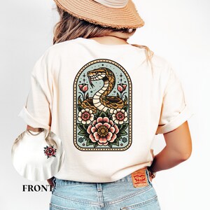 May include: A white t-shirt with a vintage-style illustration of a snake coiled around flowers in a blue and green arch. The design is framed with a gold border.