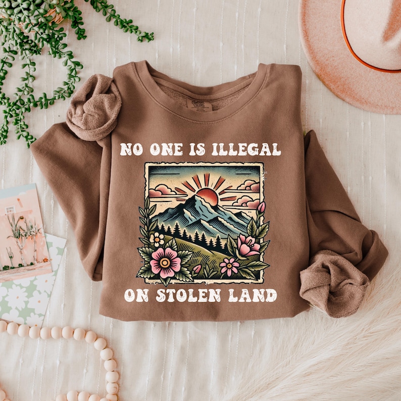 May include: Brown sweatshirt with a graphic of a mountain landscape with a red sun and the text "No one is illegal on stolen land."