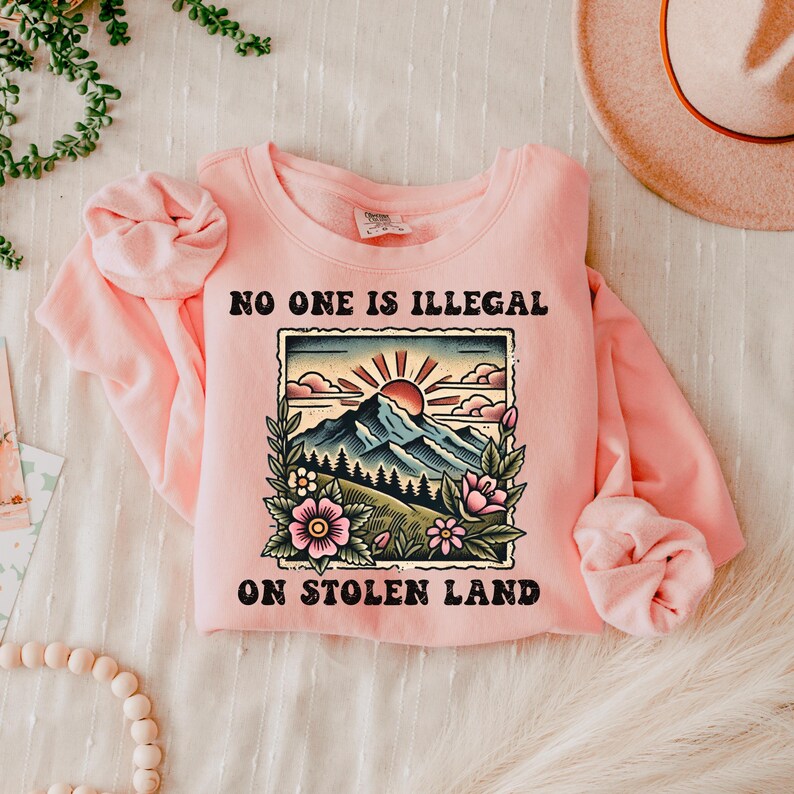 May include: Pink sweatshirt with a graphic of a mountain range, a sun, and flowers. The text "No one is illegal on stolen land" is printed on the sweatshirt.