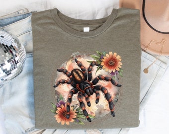 May include: A green t-shirt with a watercolor illustration of a black and orange spider surrounded by flowers. The spider has hairy legs and a black body with orange stripes.