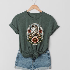 Peut inclure: Un t-shirt vert avec une illustration vintage d'un serpent enroulé autour de fleurs dans un cadre circulaire. Le serpent est brun et blanc, et les fleurs sont rouges, orange et jaunes.