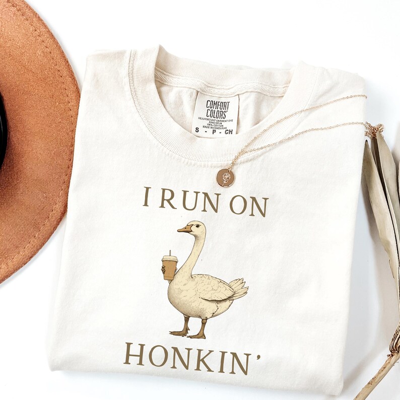 May include: A beige t-shirt with the text "I run on honkin'" printed on it. A cartoon goose is holding a cup of coffee.