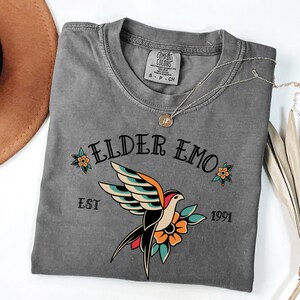 May include: A gray t-shirt with a vintage-style graphic of a bird with a flower and the text "Elder Emo Est 1991".