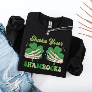 Puede incluir: Sudadera negra con un gr&aacute;fico de tr&eacute;bol verde y el texto "Shake Your Shamrocks".