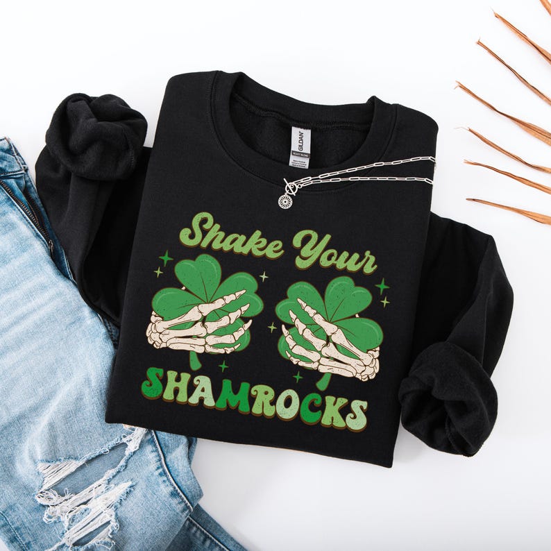 Puede incluir: Sudadera negra con un gr&aacute;fico de tr&eacute;bol verde y el texto "Shake Your Shamrocks".